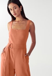 Žena s dlouhými tmavými vlasy, oblečená v oranžovém bezrukávovém overal s širokým páskem a zlatými kruhovými náušnicemi, pózuje před hladkým bílým pozadím.