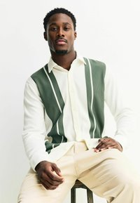 Gestreiftes Hemd mit grün-weißer gerippter Textur. Das Model trägt cremefarbene Hosen. Einfacher Kragen und sichtbare Knöpfe auf der Vorderseite.