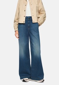 Denim wide-leg jeans, blue with slight fading, paired with a beige cropped jacket and patterned sneakers. Soft texture, relaxed fit.