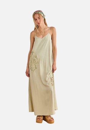 Mujer vestida con un vestido maxi sin mangas de color beige con detalles de encaje, un pañuelo en la cabeza estampado, collares en capas y sandalias de plataforma beige.