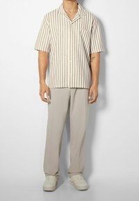 Koszula z krótkim rękawem w beżowe i bordowe pionowe paski, zapinana na guziki, z kieszenią na piersi, w zestawieniu z jasnymi spodniami i trampkami.