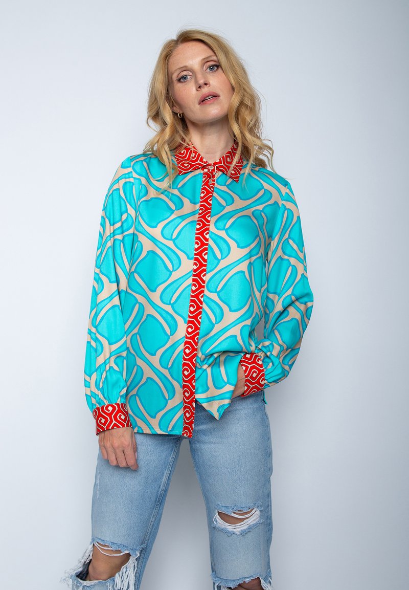 Koszula w wzory w kolorach niebieskim, beżowym i czerwonym z abstrakcyjnymi wzorami. Długie rękawy z czerwonymi mankietami, zapięcie na guziki z przodu i luźny krój.