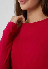 Haut rouge à manches longues en tissu doux, présentant un col rond et une coupe ajustée, montré au niveau de l'épaule avec une main touchant le tissu.