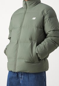 Man som bär en olivgrön puffjacka med dragkedja fram och New Balance-logotyp, med händerna i fickorna, i kombination med blå denimjeans.