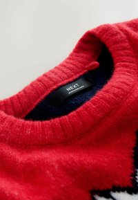 Punane kudumi svettar, millel on soonikuga ümardatud kaelus, tumesinise sisevoodriga ja must silt, millel on kirjas "NEXT". Tekstuuriga, soe kangas.