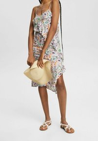 Zomerse jurk met bloemenpatroon, rimpelachtige halslijn, verstelbare spaghettibandjes en asymmetrische zoom, aangevuld met een beige geweven hoed en sandalen.