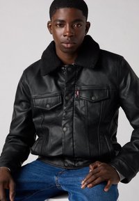 Μαύρο δερμάτινο μπουφάν με πλούσιο μαύρο γιακά, διαθέτοντας δύο εμπρόσθιες τσέπες και μεταλλικά κουμπιά. Φορεμένο πάνω από λευκό μπλουζάκι και μπλε τζιν.