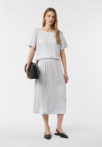 Graues Midi-Kleid mit kurzen Ärmeln, elastischer Taille, glatter Textur. Model hält eine kleine schwarze Tasche und trägt schwarze Ballerinas. Einfaches und elegantes Design.
