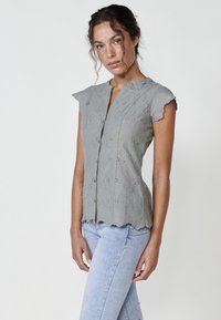 Hellgraues ärmelloses Blusenshirt mit floralem Stickereimuster, gewellten Kanten und einer knöpfbaren Vorderseite. Kombiniert mit hellblauen Skinny-Jeans.