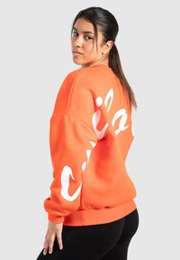 Felpa arancione con lettering bianco sul retro. Presenta un colletto a girocollo, polsini e orlo a costine. Il tessuto sembra morbido e strutturato.