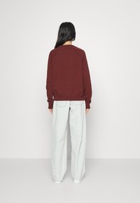 Femme se tenant de dos, portant un sweat-shirt bordeaux, un jean clair ample et des baskets blanches, sur un fond blanc uni.