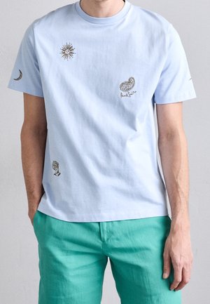 Mężczyzna w jasnoniebieskiej koszulce z grafikami słońca, księżyca, wzoru paisley i dłoni, ubrany w turkusowe spodnie, stoi z rękami w kieszeniach.