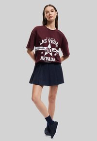 Maroon t-paita, jossa on valkoinen grafiikka, jossa lukee "LAS VEGAS NEVADA 80", yhdistettynä tummansiniseen laskostettuun hameeseen. Mallilla on tummansiniset sukat ja mustat kengät.