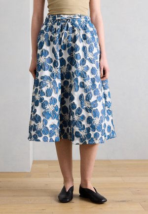 Vrouw die een knielange rok draagt met blauw-witte bloemrijke borduursels, een beige top en zwarte instap schoenen, staand op een houten vloer.