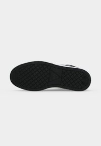 Semelle en caoutchouc noir d'une basket avec un motif de bande de roulement en forme de diamant et un logo triangulaire au centre indiquant « GUESS 7 ».