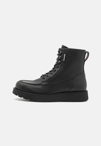 Schwarze Leder Schnürstiefelette mit dicker Sohle, Markenetikett an der Seite und Zugschlaufe an der Ferse, im Profil vor einem weißen Hintergrund dargestellt.