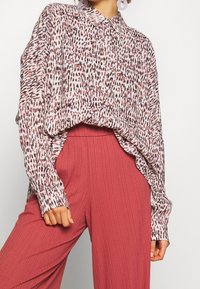 Camisa abotonada de tela ligera con un fondo blanco y manchas abstractas rojas y negras. Combinada con pantalones rosa con pliegues.