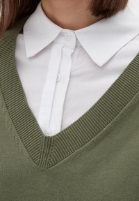 Olijfgroen gebreid sweatshirt met een V-halsontwerp, gedragen over een wit overhemd met kraag. Heeft ribbelranden en een gladde stoftextuur.