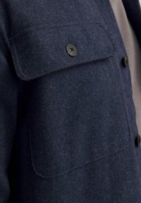 Svetle modrá bunda z vlněné směsi s texturovaným povrchem, vybavená náprsní kapsou, černým knoflíkem a jemnými stehy.