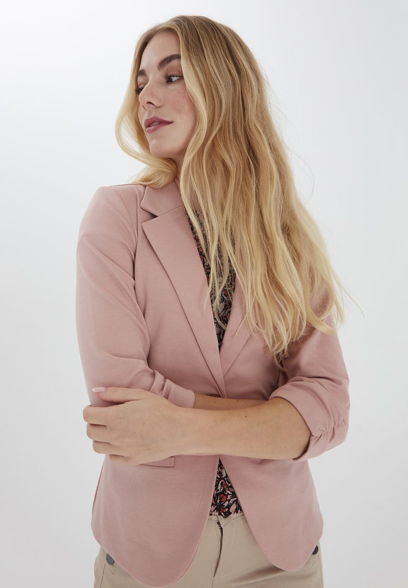 Svetle růžový blazer s propracovaným designem, klopami a řasenými rukávy do tří čtvrtin, nošený přes vzorované triko.