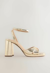 Sandale à talon haut dorée avec texture de peau de serpent, bride de cheville avec boucle, talon bloc, et brides croisées sur les orteils.