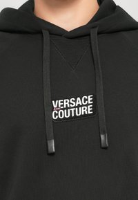 Čierna mikina s kapucňou, ktorá má na prednej strane logo nášivku s textom "VERSACE JEANS COUTURE" v bielej a ružovej farbe, a čierne šnúrky s plastovými koncami.