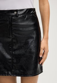 Zwarte leren minirok met een glanzende afwerking, voorzien van een sluiting aan de voorkant en zijnaad. Soepele textuur met subtiele kreukdetails.