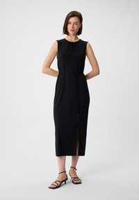 Schwarzes ärmelloses Kleid mit Rundhalsausschnitt, taillierter Taille und seitlichem Schlitz. Glatte Textur. Kombiniert mit schwarzen Riemchenabsätzen.