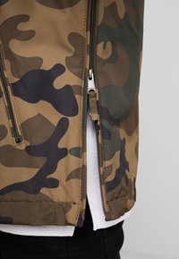 Chaqueta de camuflaje en tonos marrones y verdes, con cremalleras laterales, un forro interior suave y un corte estándar con una textura lisa.