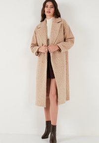 Manteau beige oversize en tissu texturé, avec des revers à cran et des boutons double-breasted, porté sur un col roulé crème et une jupe sombre.