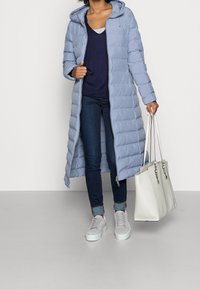 Abrigo acolchado largo azul claro con capucha, cierre frontal con cremallera y diseño acolchado. Combinado con vaqueros oscuros y zapatillas grises, llevando un bolso blanco.