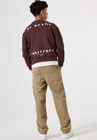 Sudadera marrón con el texto blanco "BE READY CHALLENGE" en la espalda, combinada con pantalones de pana beige y zapatillas blancas.
