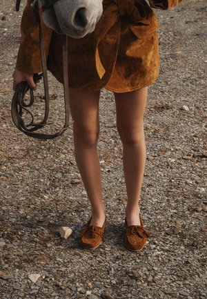 Persona con falda de ante marrón y mocasines sosteniendo las riendas cerca del hocico blanco de un caballo que está sobre un terreno rocoso.