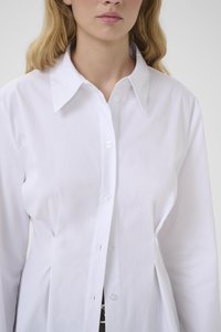 Woman with blonde hair wearing a fitted white button-up shirt with a wide collar against a plain background.