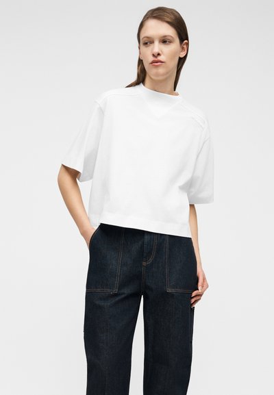 Νεαρή γυναίκα με καστανά μαλλιά φοράει άνετο λευκό κοντομάνικο μπλουζάκι και σκούρο μπλε τζιν παντελόνι, στέκεται με τα χέρια στις τσέπες.