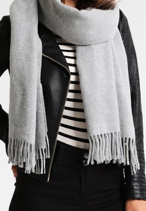 Femme portant une écharpe grise claire à franges sur une veste en cuir noire et un haut rayé noir et blanc.