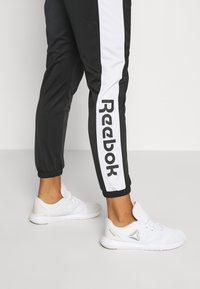 Personne portant un pantalon de survêtement Reebok noir et blanc ainsi que des chaussures de sport blanches Reebok, debout sur un sol gris devant un fond blanc.