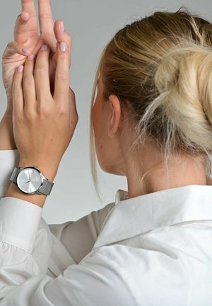 Silberne Armbanduhr mit einem Netzwerkarmband, ausgestattet mit einem runden weißen Zifferblatt mit minimalen Markierungen, getragen an einer Hand, die vor einem weißen Hemd erhoben ist.