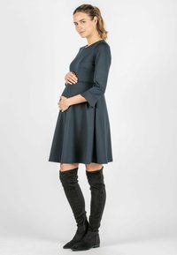 Dunkelgrünes, knielanges Kleid mit dreiviertelärmel, tailliertem Oberteil und weit schwingendem Rock. Kombiniert mit schulterlangen schwarzen Wildlederstiefeln.