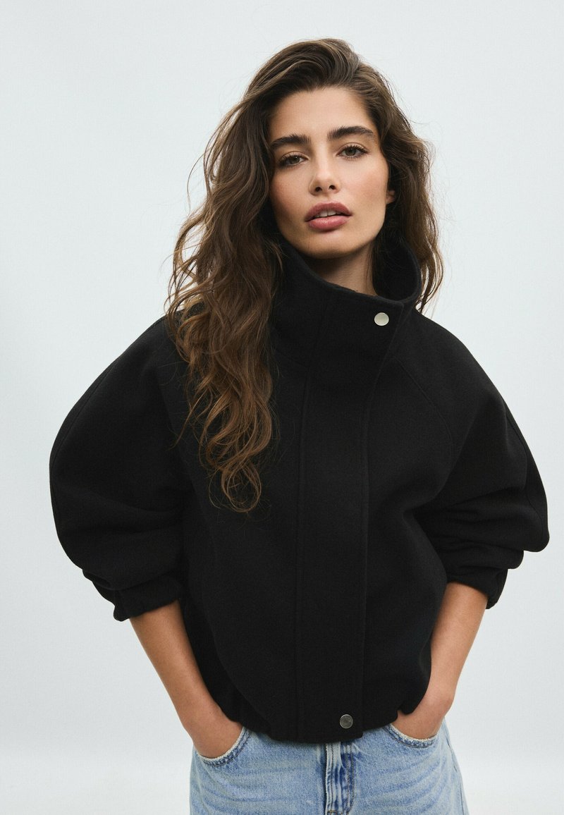 Femme aux longs cheveux ondulés portant une veste noire à col montant et un jean bleu clair, debout les mains dans les poches devant un fond clair uni.