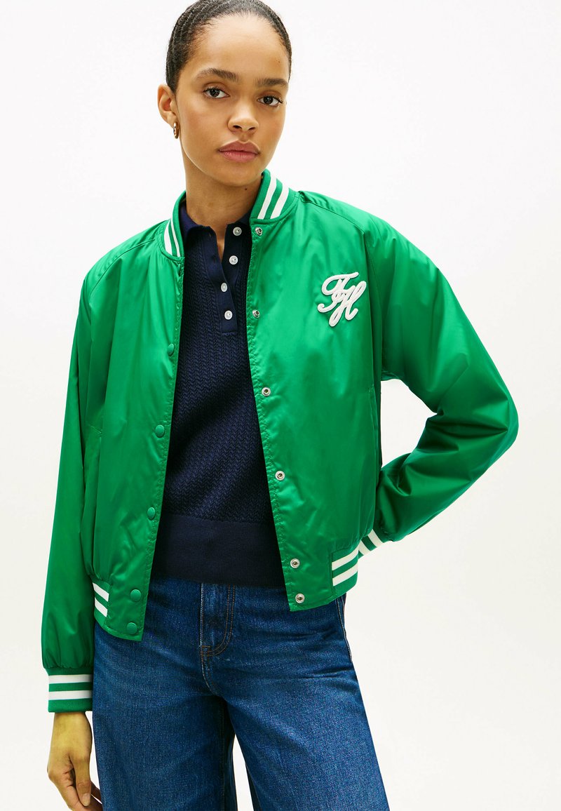Femme portant une veste universitaire vert vif avec des bandes blanches et un logo, un polo bleu marine et un jean bleu sur un fond uni.