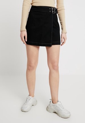 Woman wearing a black belted wrap skirt, beige long-sleeve top, and white chunky sneakers, standing against a plain background.