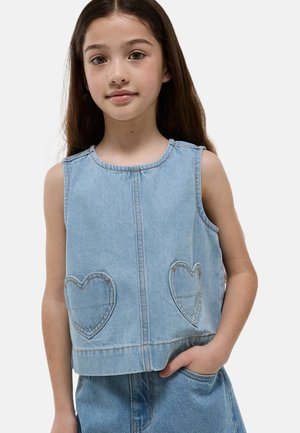Jeune fille aux longs cheveux bruns portant un haut en denim bleu clair sans manches avec des poches en forme de cœur et un bas en denim assorti.