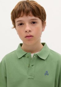 T-shirt polo vert en tissu texturé. Il possède un col à boutons et un petit emblème de crâne et os croisés bleu sur la poitrine gauche.