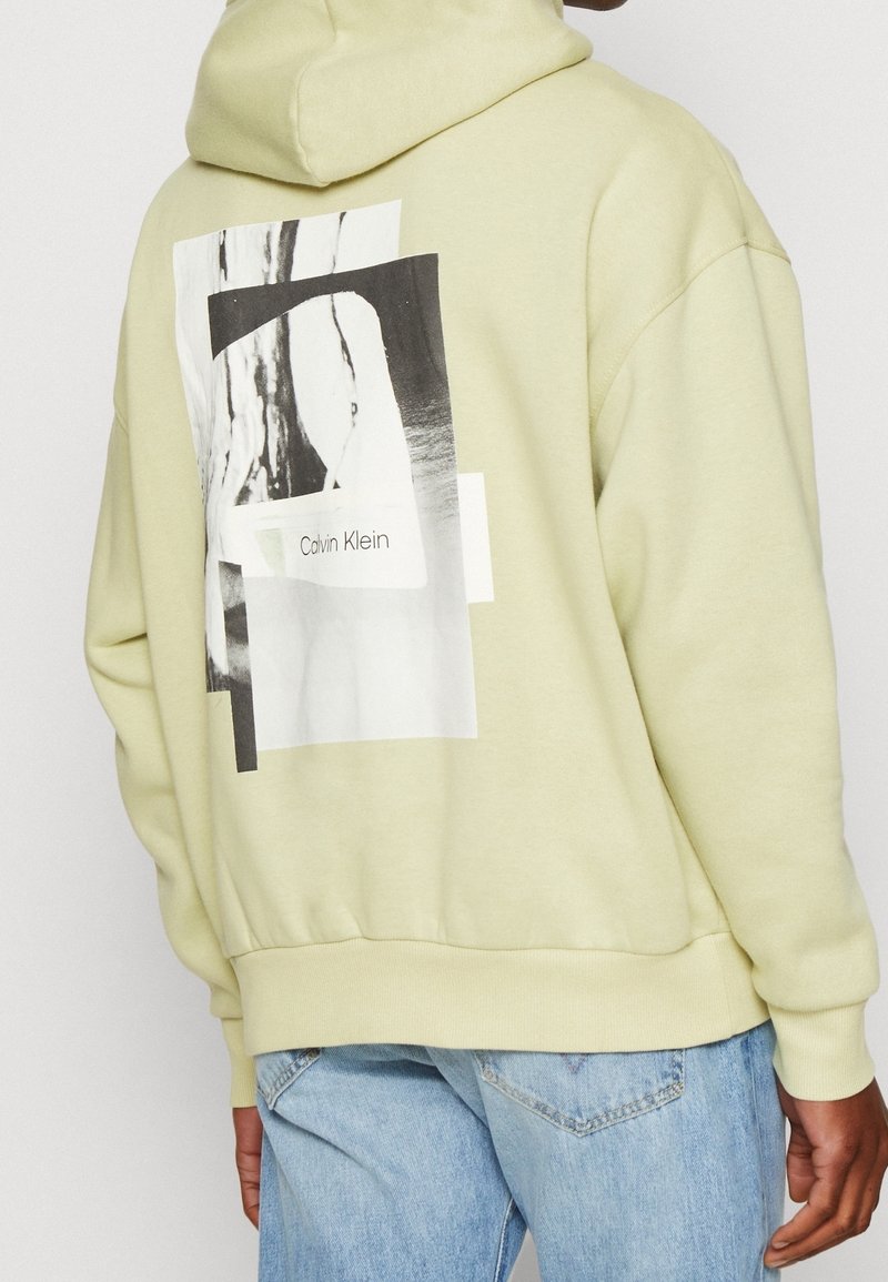Světle žlutá mikina s kapucí s grafickým designem na zádech, který obsahuje černobílé elementy a text "Calvin Klein." Měkký, uvolněný střih.