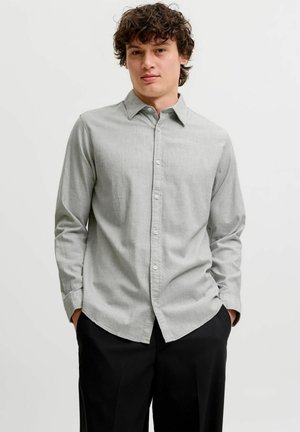 Jeune homme aux cheveux bruns bouclés portant une chemise gris clair boutonnée et un pantalon noir, les mains dans les poches, sur un fond uni.