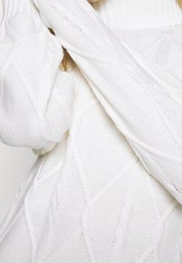 Bílý pletený svetr se strukturovaným vzorem kabelu, oversized rukávy a žebrované detaily kolem výstřihu a manžet.