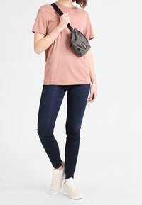 Světle růžové tričko s krátkým rukávem, tmavě modré skinny džíny a bílé tenisky. Černá a šedá crossbody taška s detaily na stahování.