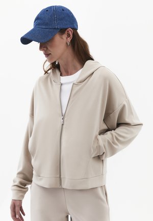 Vrouw draagt een beige zip-up hoodie en bijpassende broek met een wit shirt en een blauwe baseballpet, handen in de zakken.