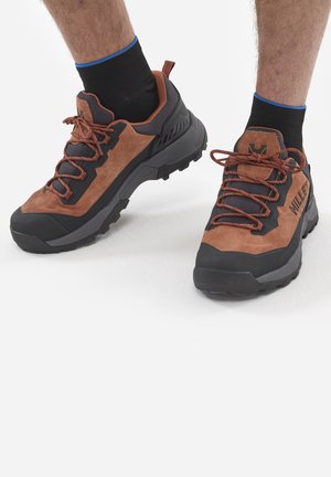 Chaussures de trail avec une tige en suédine marron et synthétique noire, capuchon en caoutchouc sur les orteils, lacets marron et semelle noire texturée. Chaussettes noires avec des accents bleus.